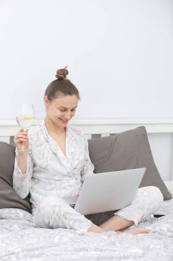 Genç bir kadın laptopunu yatakta dinlenirken ve şarap içerken kullanıyor. Evde kalmak için. Çevrimiçi çalışma. Uzak çalışma.