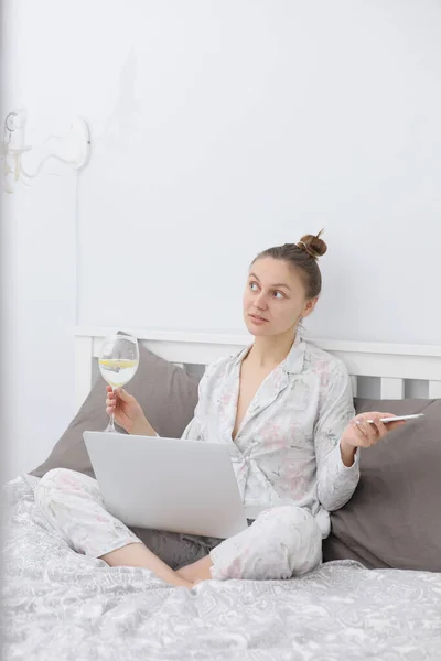 Mujer joven que usa su computadora portátil mientras se relaja en la cama y bebe vino. Quedarme en casa. Estudio en línea. Trabajo remoto. — Foto de Stock