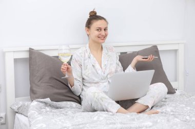 Genç bir kadın laptopunu yatakta dinlenirken ve şarap içerken kullanıyor. Evde kalmak için. Çevrimiçi çalışma. Uzak çalışma.