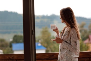 Genç bir bayan, sabahleyin otelin balkonunda duş aldıktan sonra ayakta duruyor. Elinde bir fincan kahve ya da çay tutuyordu. Doğa ormanlarının ve dağların dışına bakıyorum.