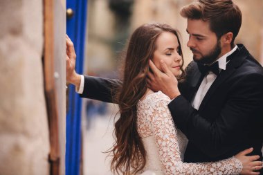 Eski şehirde düğün çifti sarılıyor. Arka planda antik kentte mavi renkli kapılar ve kafe var. Beyaz uzun elbiseli şık gelin ve takım elbiseli ve papyonlu damat. düğün günü.