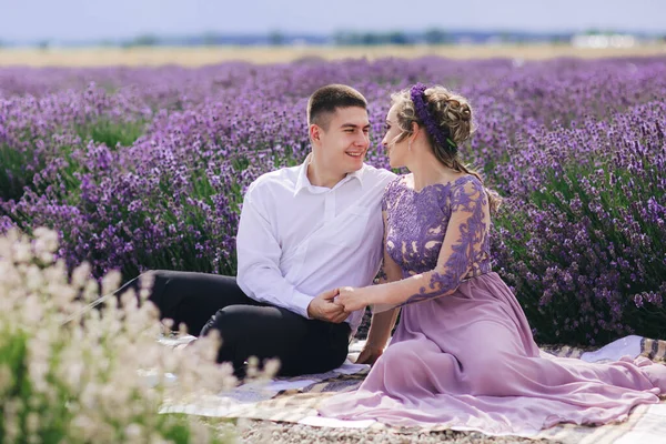 在夏天的阴冷的日子里 年轻夫妇拥抱在一起 坐在薰衣草地里 穿着华丽的紫色衣服 头戴淡紫色花环的女孩 — 图库照片