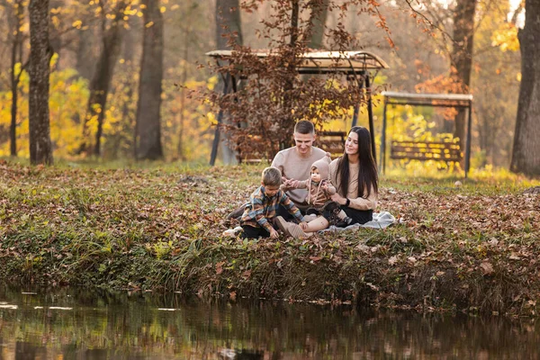 young dad and mom with baby girl and little son having fun on blanket near pond in autumn park on sunny day. happy family concept. mother's, father's, baby's day