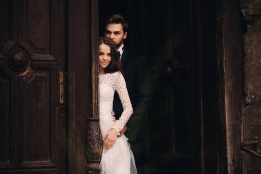 Güzel yeni evliler antik kapının yanında sarılıyorlar. Şık bir damadın düğün portresi ve Avrupa 'da eski bir evin yakınında genç bir gelin..