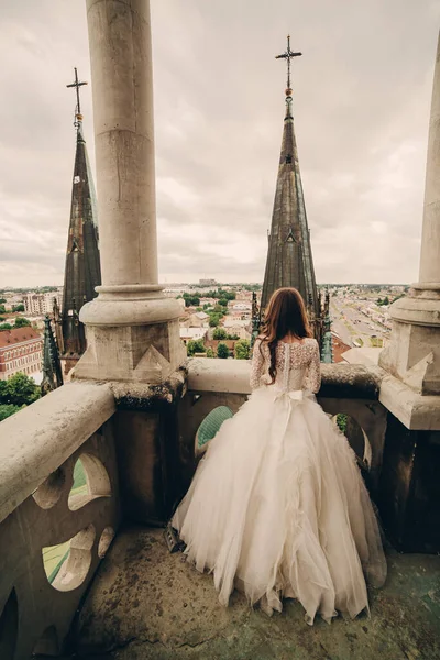 Feliz recién casada. hermosa novia en vestido blanco de lujo largo en el balcón de la antigua catedral gótica con vistas panorámicas a la ciudad —  Fotos de Stock