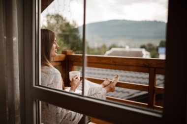 Genç bir bayan, sabahleyin otelin balkonunda duş aldıktan sonra ayakta duruyor. Elinde bir fincan kahve ya da çay tutuyordu. Doğa ormanlarının ve dağların dışına bakıyorum.