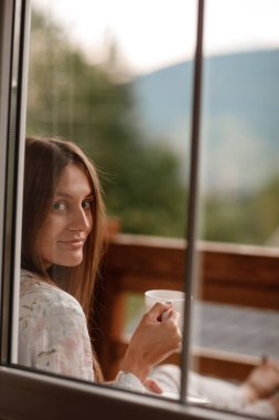 Genç bir bayan, sabahleyin otelin balkonunda duş aldıktan sonra ayakta duruyor. Elinde bir fincan kahve ya da çay tutuyordu. Doğa ormanlarının ve dağların dışına bakıyorum.