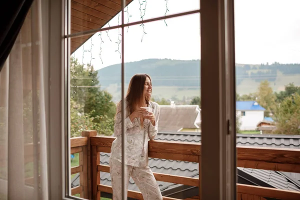 Jonge vrouw staande na een douche in de ochtend op het balkon van het hotel. met een kop koffie of thee in haar handen. Buiten het natuurbos en de bergen kijken — Stockfoto