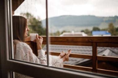 Genç bir bayan, sabahleyin otelin balkonunda duş aldıktan sonra ayakta duruyor. Elinde bir fincan kahve ya da çay tutuyordu. Doğa ormanlarının ve dağların dışına bakıyorum.