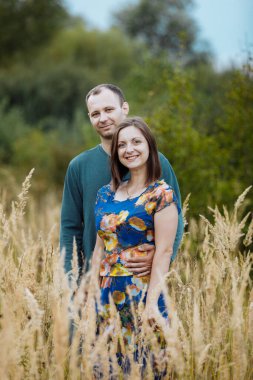 Genç karı koca el ele tutuşan çimlerle bir tarlada kucaklaşıyorlar. Mutlu aile günleri..