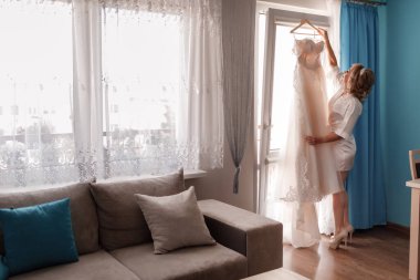 Gelin sabahı hazırlığı. İpek bornozlu mutlu genç kadın pencerenin yanında dururken gelinliğine dokunuyor..