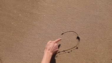 Kız plajda bir kalp çiziyor. Deniz suyu, kumlu bir sahildeki çizimleri temizler..