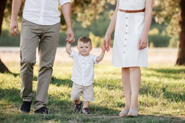 Yeni yürümeye başlayan çocuğa bak. Anne, baba el ele tutuşun, oğul parkta yürüyün. Genç bir aile tatilde, dışarıda birlikte vakit geçiriyor. Aile yaz tatili kavramı. seçici odak.