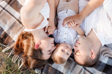 Anne, baba ve küçük oğlu yaz parkında battaniyenin üzerinde yatıyor. Yaz tatili kavramı. Anneler günü, babalar günü, bebekler günü. Aile doğada birlikte vakit geçiriyor. Aile görünüşü.