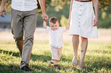 Yeni yürümeye başlayan çocuğa bak. Anne, baba el ele tutuşun, oğul parkta yürüyün. Genç bir aile tatilde, dışarıda birlikte vakit geçiriyor. Aile yaz tatili kavramı. seçici odak.