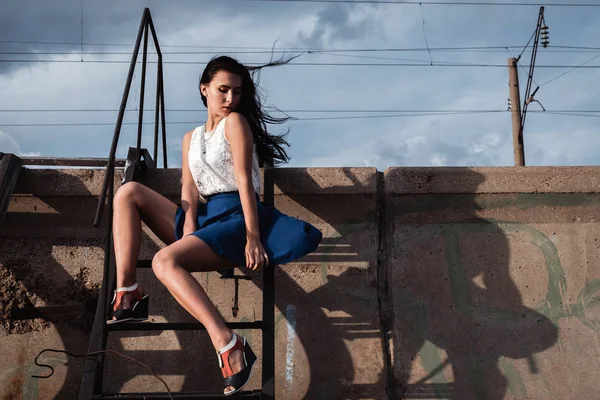 Girl in skirt sitting on the iron stairs bent legs. The wind is blowing through her hair. Behind her concrete, wires and clouds