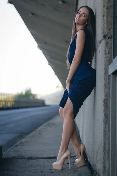 attractive woman in a long blue dress stands on her heels along a concrete wall. She has long hair, a belt and a pretty face.