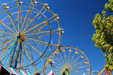 Bir tarafta ve renkli çadır pavyonlar alt ağaçların arkasında parlak mavi gökyüzü ile iki dönme dolap
