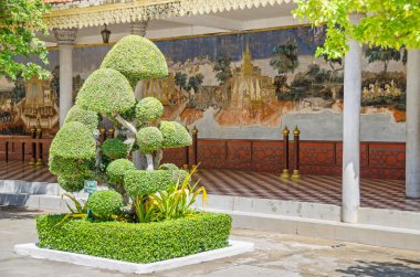 Phnom Penh, Cambodia - April 8, 2018: Reamker fresco murals of the Open-Air-Pavillon or Reamker gallery in a compound of The Silver Pagoda with a scissored Siamese rough bush or a toothbrush tree. clipart