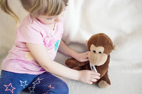 Baby with toy monkey. Little girl measure temperature for toy. Children\'s toy with medical thermometer. Beautiful little girl, long eyelashes, white hair, short tails.