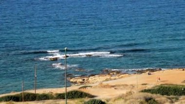 Yükseklikten deniz manzarası, parlak güneşli bir sabah, güzel bir mavi deniz, küçük bir sörf, parlak bir kumlu plaj. İki yaşlı insan sahil boyunca yürür