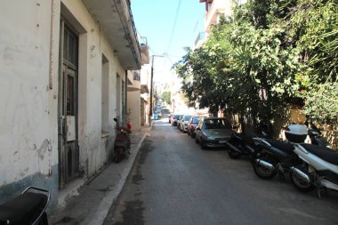 Agios Nikolaos, Girit, Yunanistan, 10/14/2015 Açıklayıcı Editoryal Evlerin ve ağaçların gölgesinde dar bir sokak, boyunca park edilmiş arabalar.
