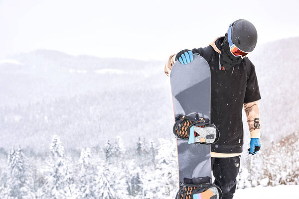 Male snowboarder looking on his snowboard on the hilltop with mountains and forrest on the background                               