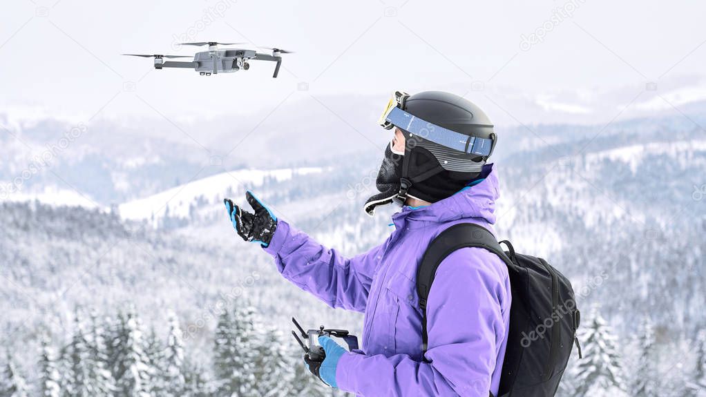 Male snowboarder in the violet jacket and black helmet launching a flying drone with a remote controller in his hand on winter mountain                               