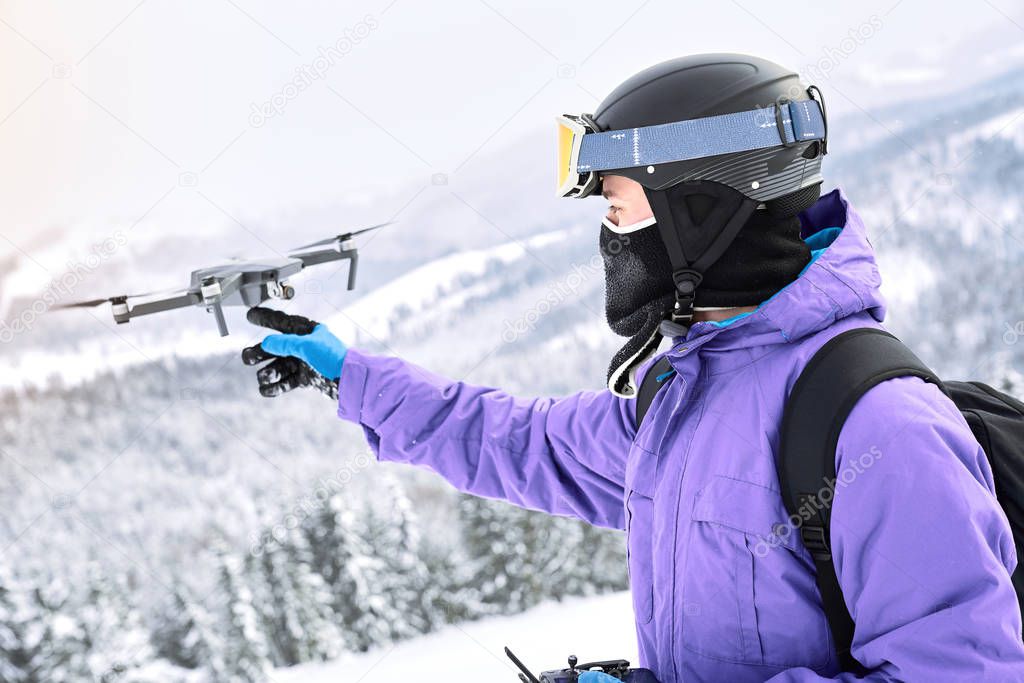 Male snowboarder with equipment and black backpack is launching a flying drone with a remote controller in his hand on winter mountain                               