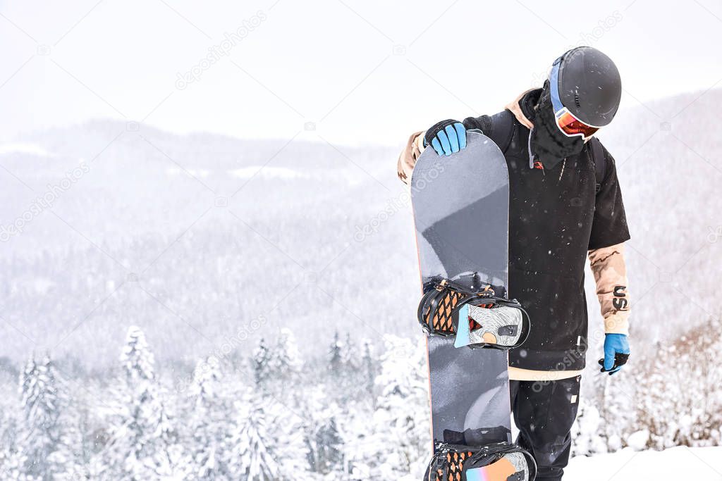 Male snowboarder looking on his snowboard on the hilltop with mountains and forrest on the background                               