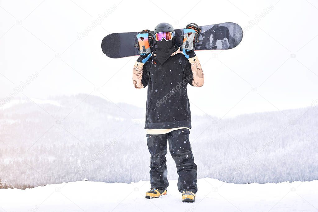 Snowboarder on the hill. Shot of the man snowboarder holding  board on his shoulders and looking front of him                               