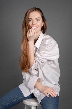 Gülümseyen ile açık kahverengi saçlı çekici kadın dikey stüdyo çekim görüntülemek ve bar tabure gri arka plan oturan fotoğraf makinesinde bak