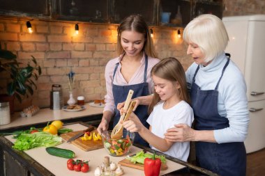 Yan görünümü büyükanne torunu salata tahta kaşıklar cam kase içinde karıştırın öğretim bir kadeh. Gülümseyen anne üzerlerine mutfakta arıyorsunuz