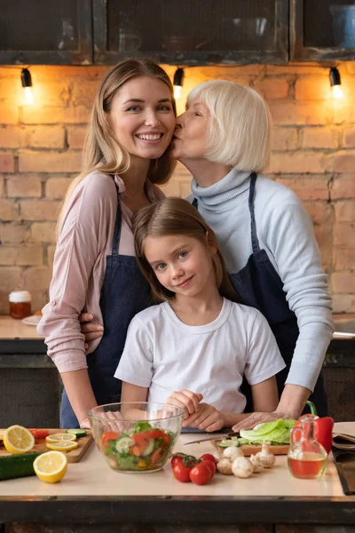 Vertical shot of elder woman that kissing and hugging her daughter in kitchen. Little cheerful girl standing near her family with look in camera