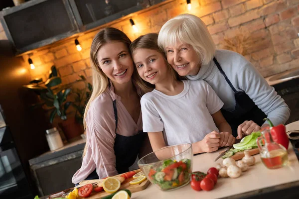 Happy family smiling and looking in camera while preparing healthy salad in kitchen. Vegetables and fruits on table