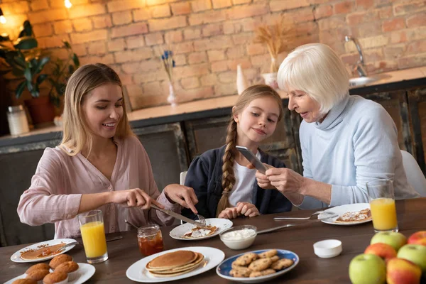 Horizontal shot of happy family at the kichen table dining. Smiling grandmother taking picture of the plate with pancakes that eating granddaughter