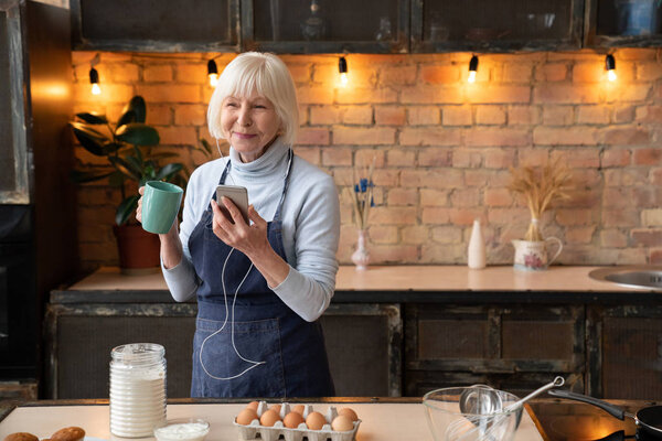 Portrait of happy mature woman chilling in kitchen while cooking. Smiling senior woman with cup of tea listening music on mobile phone in kitchen
