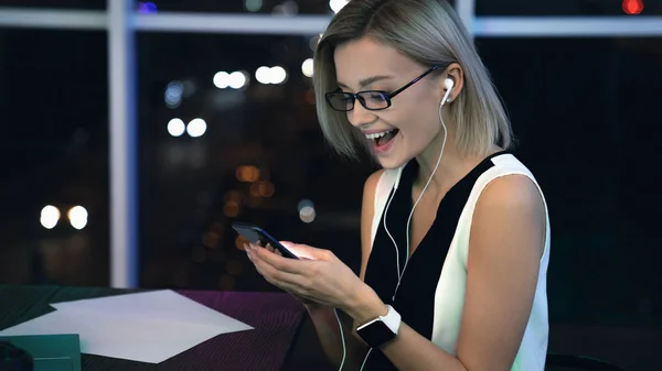 Ofiste oturan ve akıllı telefonda kısa mesaj okurken mutlu genç kadın. Ofis geç saatlerde çalışırken cep telefonları ile telefon kullanarak iş kadını. — Stok fotoğraf