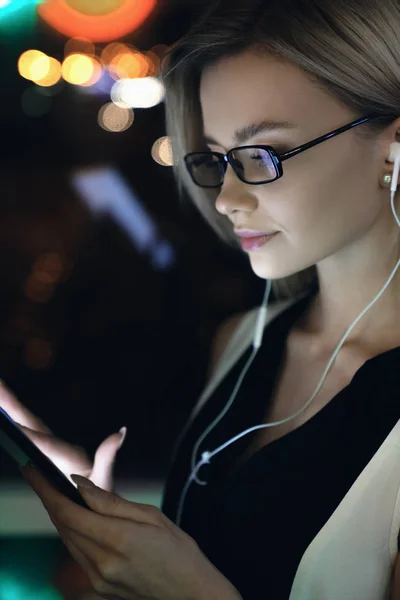 Gambar vertikal seorang wanita muda yang cantik mengirimkan surat elektronik dengan tablet di tangan dan earphone pada malam hari di kantor dengan lampu di latar belakang — Stok Foto