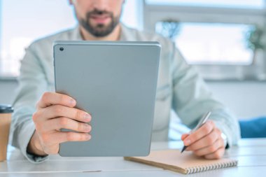 Serbest çalışan erkek yönetici bir sehpada oturan notlar yapıyor. Bir kafede dijital tablet üzerinde çalışan adam
