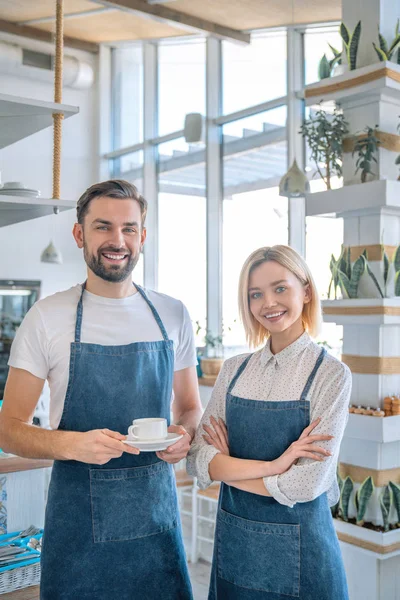 Happy coffee shop owner couple standing inside their shop. Man and woman baristas standing inside their cafe wearing apron with the man holding coffee cup — Stock Photo, Image