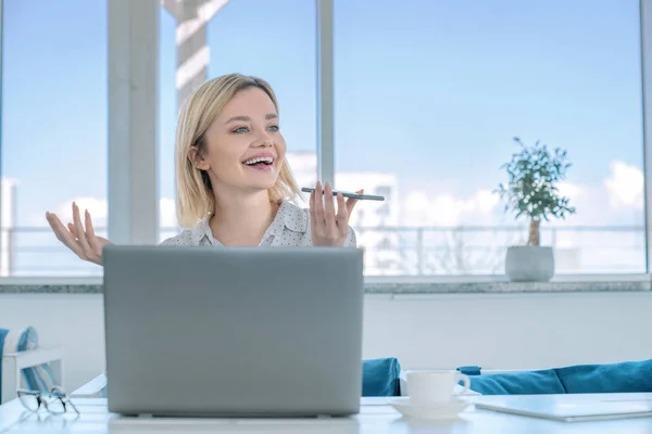 Καυκάσια επιχειρηματίας που κάθεται στο χώρο εργασίας της με φορητό υπολογιστή στο γραφείο εργασίας και συζητώντας με την αντιμετώπιση κάποιων ερωτήσεων εργασίας με ηχείο με smartphone στο χέρι με γυαλιά και κούπα καφέ σε — Φωτογραφία Αρχείου