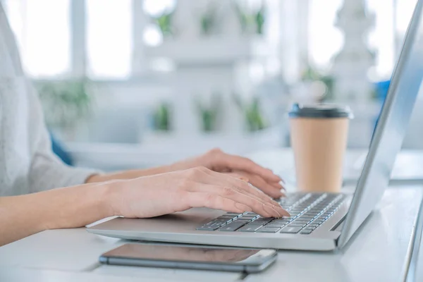 Nahaufnahme einer Geschäftsfrau, die am Laptop arbeitet, während sie mit einer Tasse Kaffee und einem Mobiltelefon an ihrem Arbeitsplatz sitzt — Stockfoto