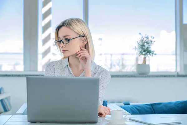 Tampilan depan foto seorang wanita muda yang bijaksana berkacamata dengan pemandangan duduk di atas meja kerja dengan laptop di atasnya di tempat kerja bersama . Stok Foto