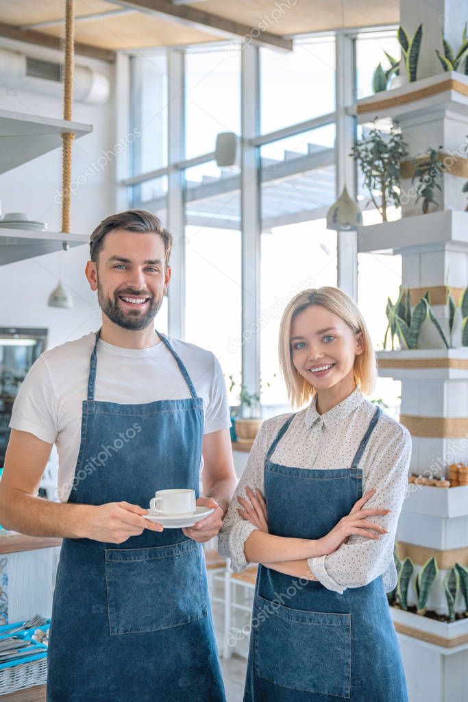 Happy coffee shop owner couple standing inside their shop. Man and woman baristas standing inside their cafe wearing apron with the man holding coffee cup