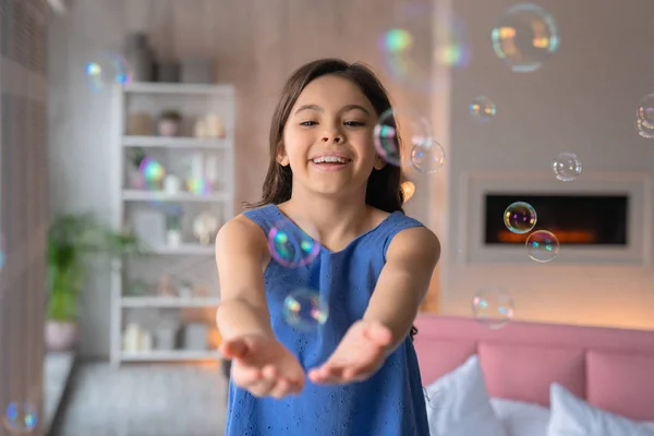 Vista frontal tiro de menina bonito brincando com o ventilador de bolhas em casa com lareira no fundo — Fotografia de Stock