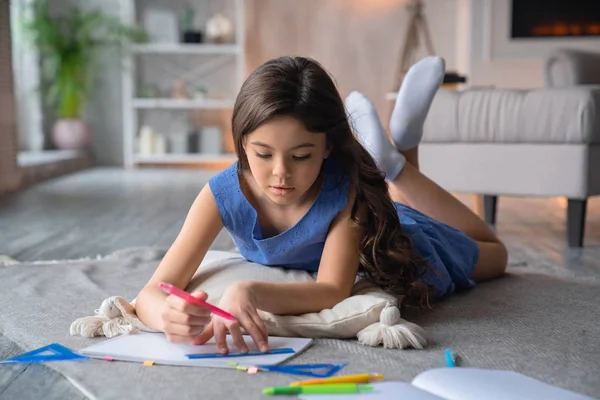 Маленькая девочка делает домашнее задание дома, лежа на полу — стоковое фото
