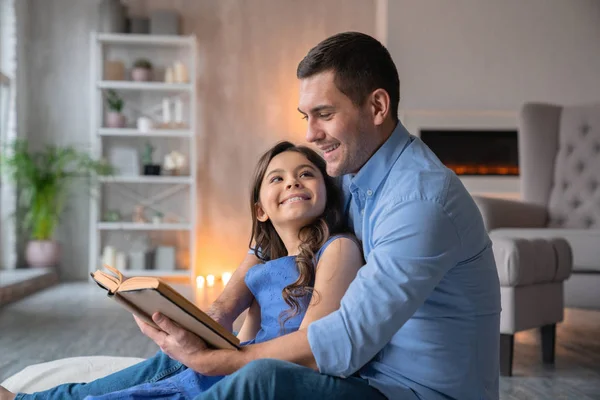 Tiro horizontal de padre sonriente ese libro de lectura con su linda hijita en casa. Pequeña chica alegre mira a su padre — Foto de Stock