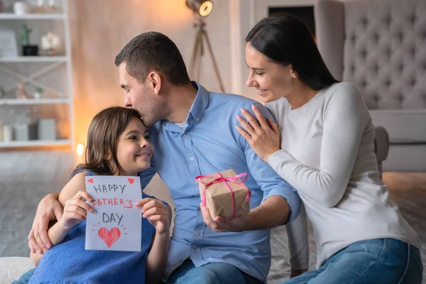 父の日おめでとう!父の日を祝う幸せな家族の正面図。贈り物を持つ若い男は、妻が床に座って夫を抱きしめている。お父さんのためのプレゼントカードを持つ小さな女の子 — ストック写真