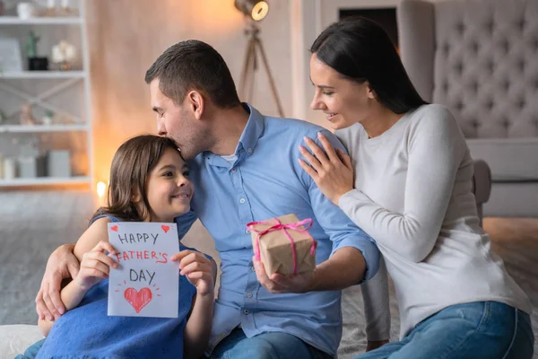 Szczęśliwy dzień ojca! Widok z przodu strzał szczęśliwej rodziny obchodzi dzień ojców. Młody mężczyzna z darem w rękach jego żona siedzi na podłodze i przytulanie męża. Mała dziewczynka z obecną kartą dla taty — Zdjęcie stockowe